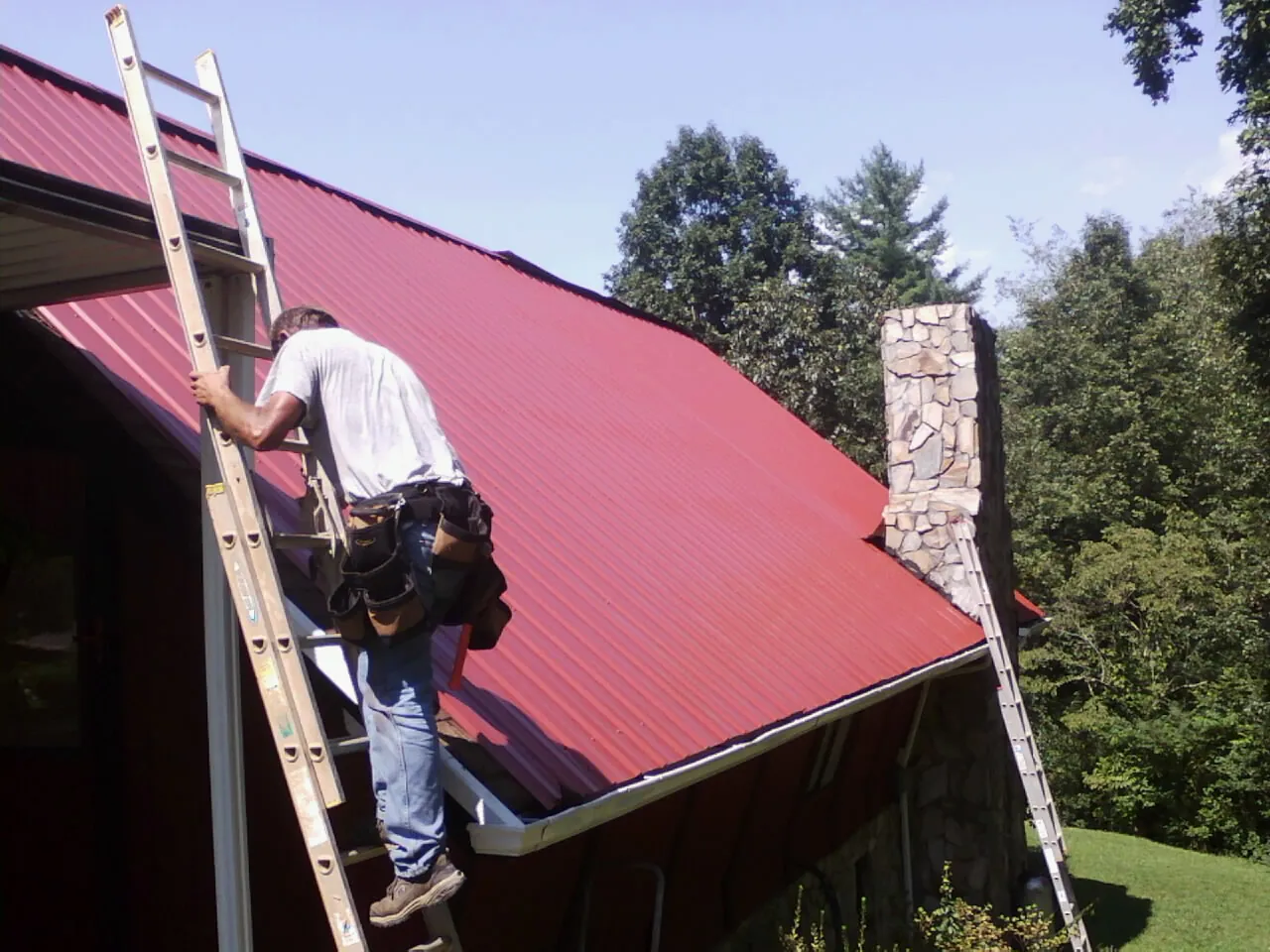 Roofer on a ladder beside a red metal roof and stone chimney — Soffit & Fascia Repair by Roofing Brothers in Leonia
